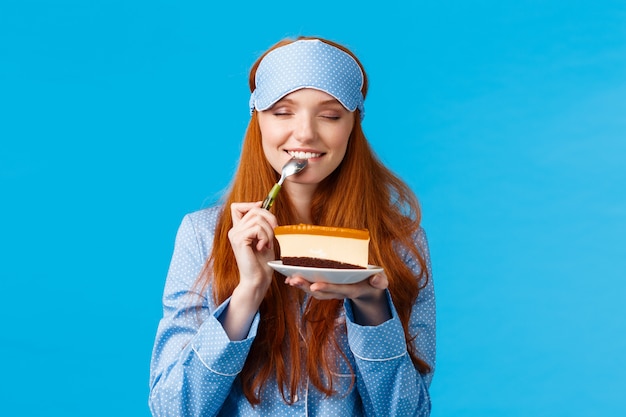 Satisfacción de la muchacha que come el postre delicioso dulce en mañana. Atractiva mujer linda con el pelo rojo en ropa de dormir y antifaz para dormir, ojos cerrados mordiendo sabroso pastel, sosteniendo los ojos cerrados con cuchara