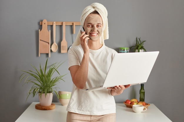Satisfecha y exitosa gerente en línea de una joven posando en la cocina con una toalla en la cabeza combina procedimientos de cuidado de la piel y trabajo en línea sosteniendo una computadora portátil y hablando por teléfono