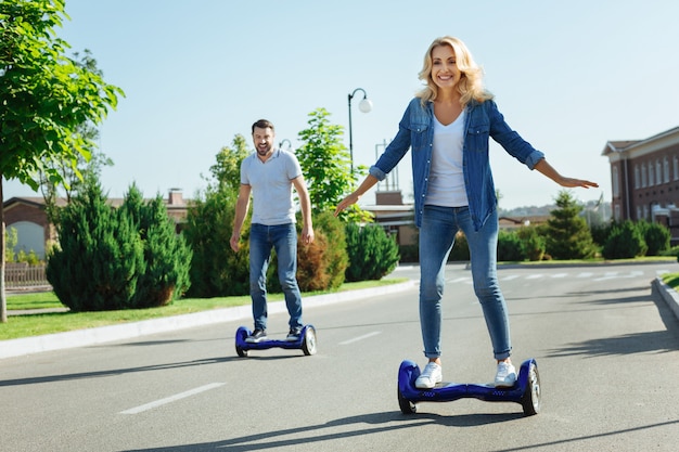 Satisfecho con la compra. Marido y mujer jovenes alegres montando patinetas y sonriendo felizmente, contentos con su nueva compra