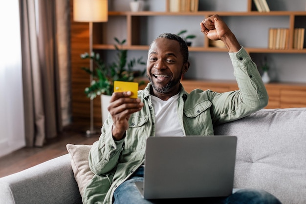 Satisfecho y emocionado hombre afroamericano maduro se regocija de una gran compra y devolución de dinero con una computadora portátil y una tarjeta de crédito
