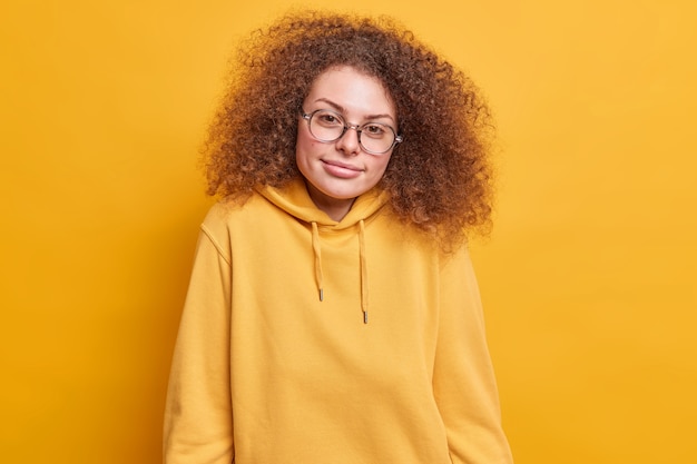 Satisfecho estudiante bonita de pelo rizado con expresión seria lleva gafas transprarent y sudadera casual aislado sobre la pared amarilla. Concepto de expresiones de rostro humano de personas