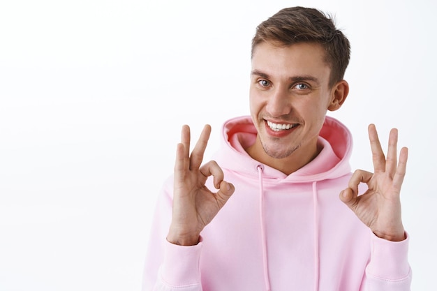 Satisfecho, guapo joven rubio con sudadera con capucha rosa, absolutamente como su gran idea, muestre un gesto correcto, garantice la mejor calidad, recomiende el producto, esté de pie en una pared blanca satisfecha, sonriendo