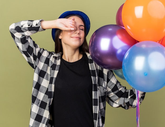 Foto satisfecho con los ojos cerrados joven hermosa niña con sombrero azul sosteniendo globos ojo cubierto con la mano