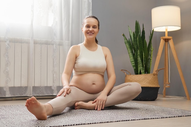 Foto satisfeita, encantada, grávida, vestindo top bege e leggings no interior leve da sala de estar, com expressão facial feliz, com gravidez em casa.