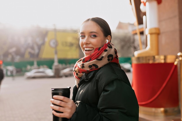 Satisfeita mulher adorável com cabelos escuros e maquiagem brilhante usando cachecol brilhante e jaqueta escura se virar para a câmera à luz do sol com café enquanto caminhava na cidade