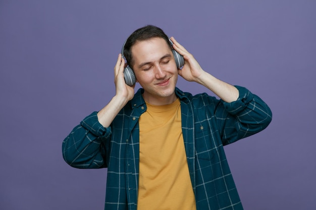 Satisfeito jovem estudante do sexo masculino usando fones de ouvido, mantendo as mãos em fones de ouvido, ouvindo música com os olhos fechados, isolados no fundo roxo