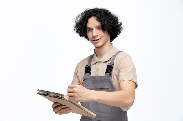 Satisfeito jovem trabalhador da construção civil vestindo uniforme segurando lápis e bloco de notas tomando notas de trabalho olhando para a câmera isolada no fundo branco