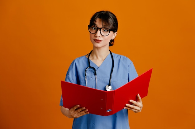 Satisfeito segurando a pasta jovem médica vestindo uniforme quinto estetoscópio isolado em fundo laranja