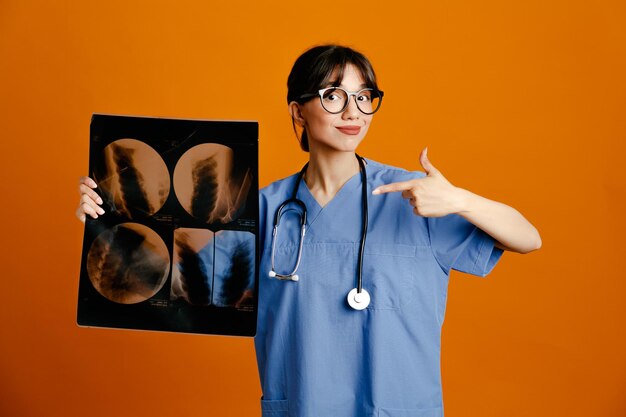 Satisfeito segurando e apontando para a jovem médica de raio-x usando estetoscópio uniforme de quinto isolado em fundo laranja