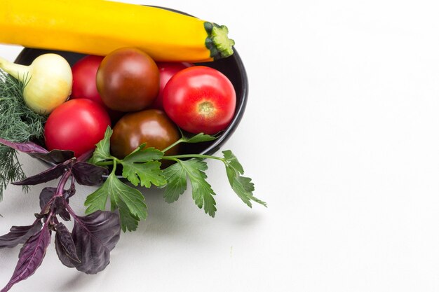 Satz Gemüse und Zweige Basilikum und Petersilie in schwarzer Schüssel. Gelbe Zucchini und rote Tomaten. Weißer Hintergrund. Platz kopieren. Ansicht von oben