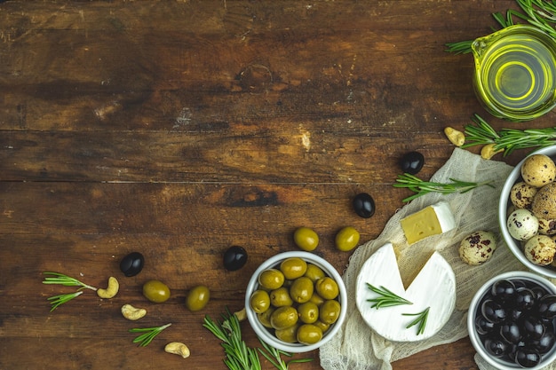 Satz Käse-Camembert, schwarze und grüne Oliven, Wachteleier auf Tellern, Olivenöl und Rosmarin-Cashewnüsse auf altem dunklen Holztischhintergrund, Kopierraum von oben