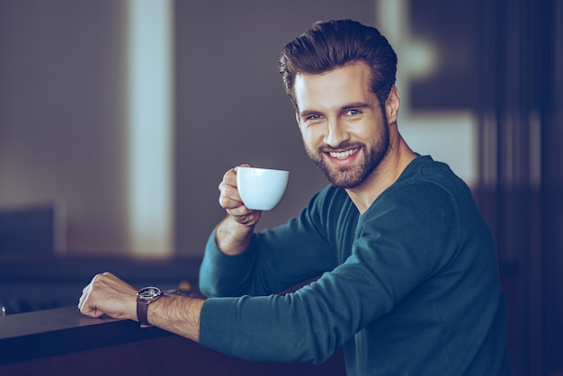 Saudação! vista lateral de um jovem bonito segurando a xícara de café e olhando para a câmera com um sorriso enquanto está sentado no balcão do bar