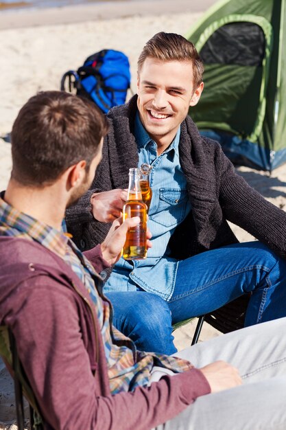 Saudações à amizade! Vista superior de dois belos rapazes comemorando com cerveja e sorrindo enquanto estão sentados perto da tenda e na areia