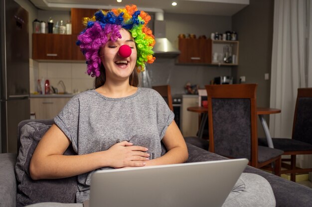 Foto saudações de feriado online, jovem se comunica em redes sociais com laptop, garota sentada em casa na cadeira com laptop, palhaço com laptop em casa, rir