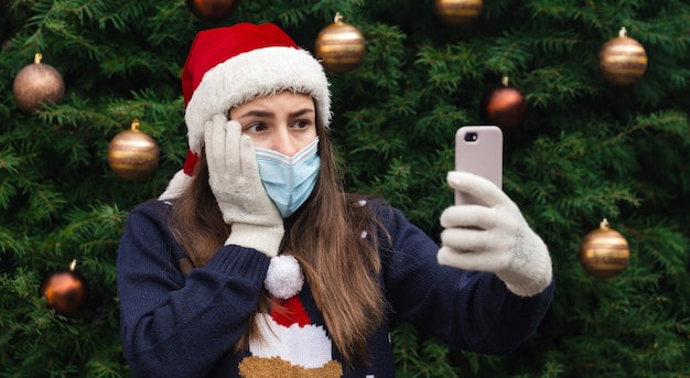 Saudações online de natal. feche o retrato de uma mulher com um chapéu de papai noel e uma máscara médica com emoção. no contexto de uma árvore de natal. pandemia do coronavírus