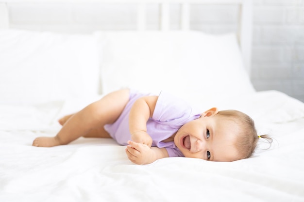 Saudável menina fofa criança em um bodysuit encontra-se de costas na cama na cama branca, olha para a câmera. O conceito de produtos para bebês.