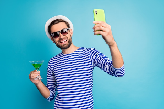 Saúde! foto de um viajante descolado tomando blog telefone selfies beber coquetel verde resort all inclusive usar óculos de sol listrado boné de camisa de marinheiro isolado cor azul