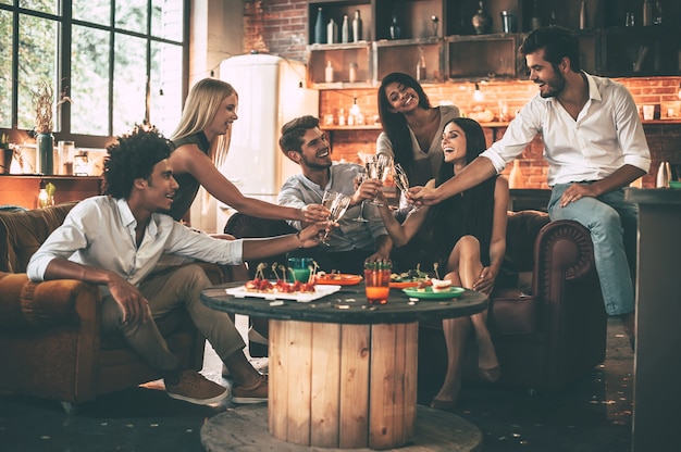 Saúde! Grupo de jovens alegres desfrutando de comidas e bebidas enquanto passam bons momentos em cadeiras confortáveis na cozinha.