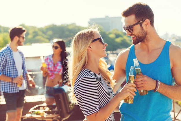 Saúde para nós! Casal jovem sorridente, tilintando de copos com cerveja e olhando um para o outro, enquanto duas pessoas fazem churrasco ao fundo