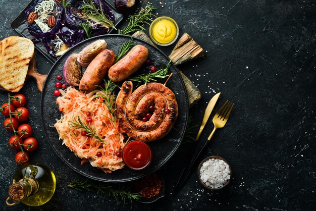 Sauerkraut und gegrillte Würstchen mit Rosmarin und Preiselbeeren auf einem schwarzen Teller Traditionelles deutsches Gericht Draufsicht auf schwarzem Steinhintergrund