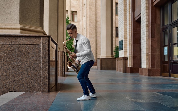 Saxofonista masculino toca el saxofón en la calle, talento joven. Músico con saxo al aire libre, actuación musical en la ciudad, intérprete de jazz