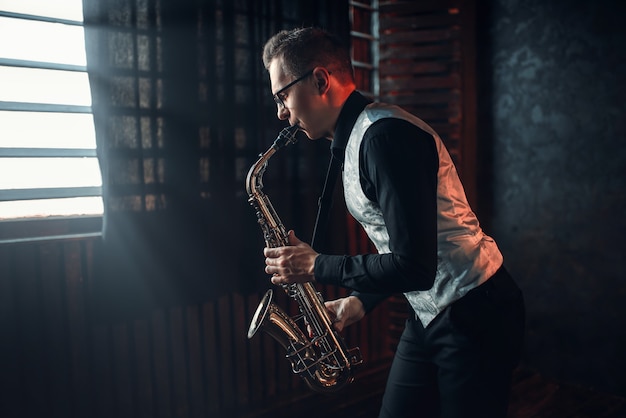 Foto saxofonista masculino tocando melodía de jazz en el saxofón contra la ventana