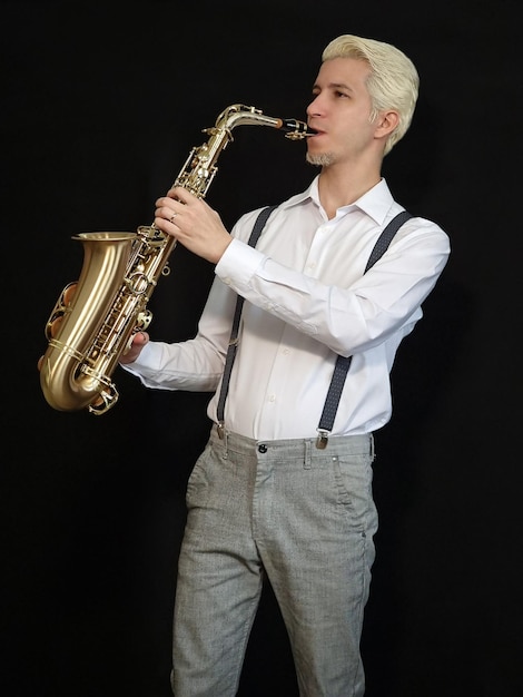 Foto saxofonista rubio con camisa blanca y tirantes grises tocando el saxofón dorado con fondo negro