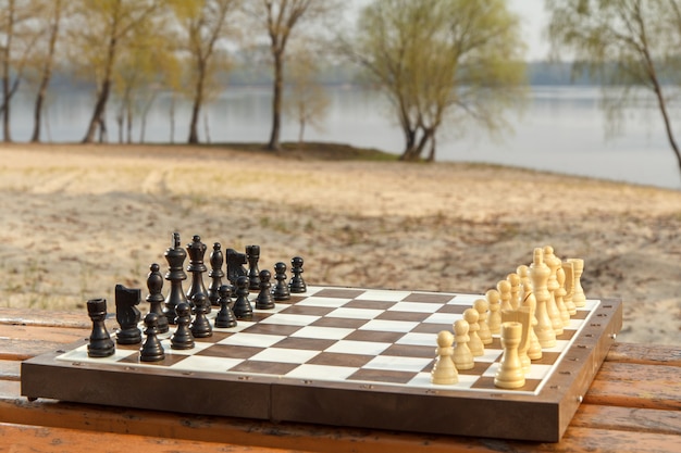 Schachbrett mit Schachfiguren auf Holzbank mit Flussuferhintergrund. Schachspiel mit Holzschachfiguren