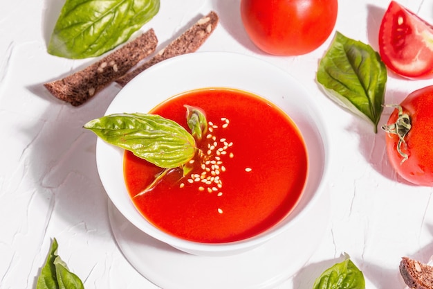 Schalen mit hausgemachter Tomatensuppe. Reifes Gemüse, frische Basilikumblätter, Grissini, aromatische Gewürze. Trendiges hartes Licht, dunkler Schatten. Weißer Kitthintergrund, flach