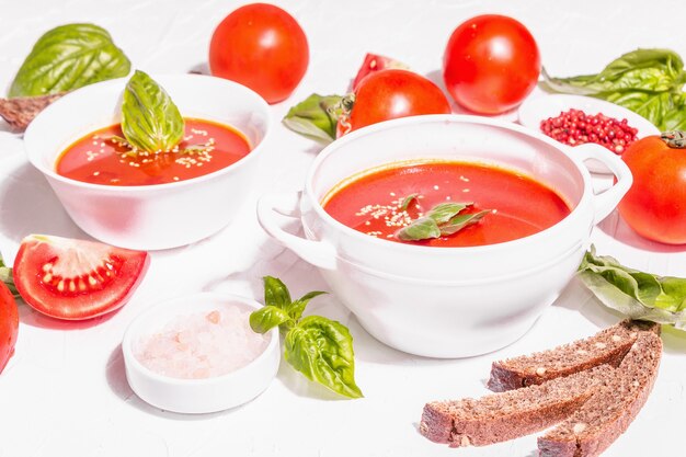 Schalen mit hausgemachter Tomatensuppe. Reifes Gemüse, frische Basilikumblätter, Grissini, aromatische Gewürze. Trendiges hartes Licht, dunkler Schatten. Weißer Kitthintergrund, flach