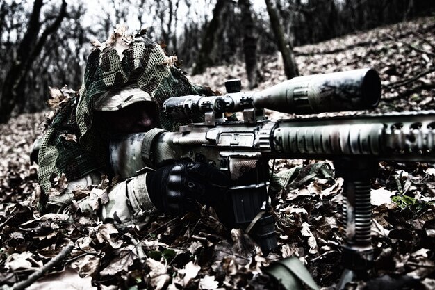 Scharfschütze der Armee-Elitetruppen, taktischer Gruppenschütze, Airsoft-Spieler, der im Wald auf dem Boden liegt, sich im Herbstlaub versteckt, sich mit einem Tarnumhang bedeckt, Ziele durch optisches Visier sucht