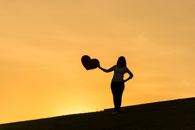 Schattenbild der asiatischen Dame stehend auf Hügel und Herz auf der Seite während der Sonnenuntergangzeit halten