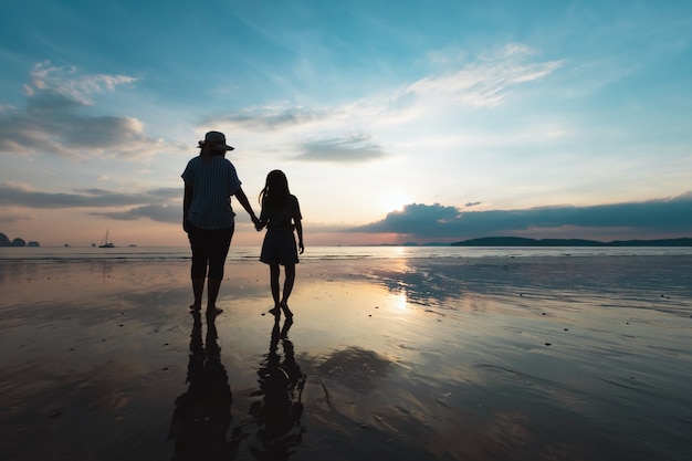Schattenbild der asiatischen Mutter und der Tochter, die Hand halten und zusammen auf dem Strand zur Sonnenuntergangszeit mit schönem Meer und Himmel gehen. Familie genießen mit Naturkonzept.