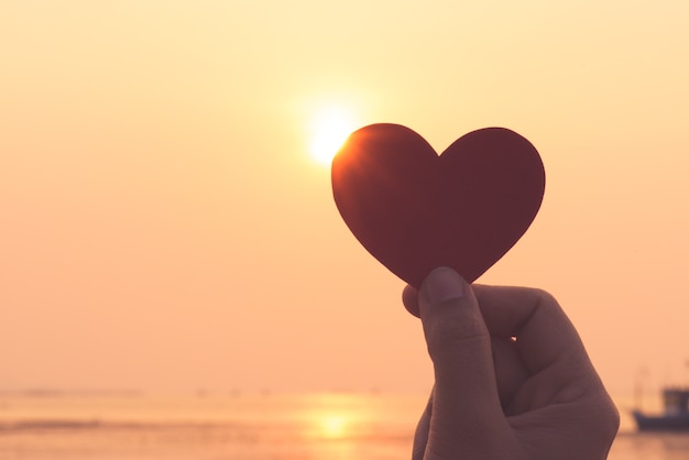 Schattenbild der Hand rotes Herz während des Sonnenunterganghintergrundes halten.