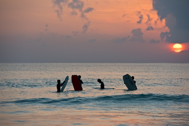 Schattenbild der Jungen, die auf Brettern im Ozeanufer während des Sonnenuntergangs surfen