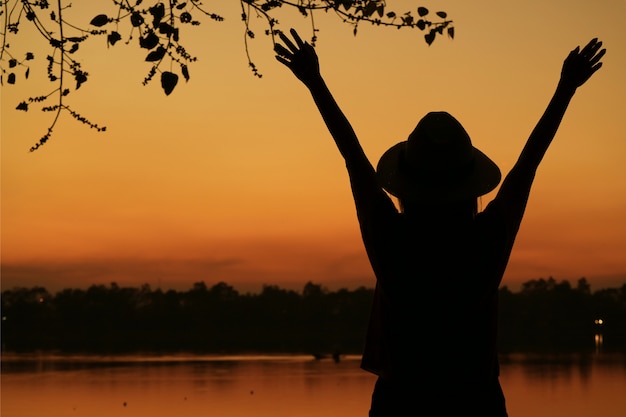 Schattenbild der jungen Frau Arme gegen schönen orange Farbsonnenunterganghimmel auf dem Seeufer anhebend