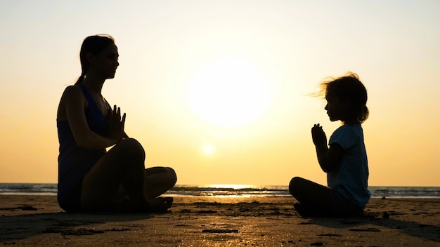 Schattenbild der mutter mit der kleinen tochter, die zusammen in der türkischen pose am strand bei sonnenuntergang meditiert