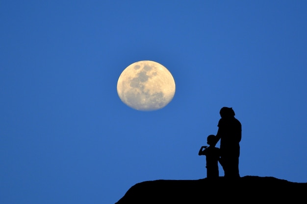 Schattenbild der Mutter und des Sohns, die auf dem Berg, den blauen Hintergrund des Mondes aufpassend stehen.