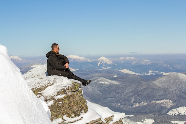 Schattenbild des alleinigen Touristen, der auf schneebedeckter Bergspitze sitzt, die Ansicht und Leistung am hellen sonnigen Wintertag genießt. Abenteuer, Aktivitäten im Freien, gesunder Lebensstil.