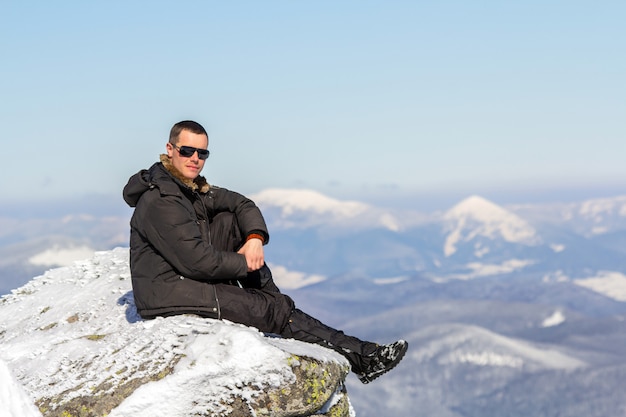 Schattenbild des alleinigen Touristen, der auf schneebedeckter Bergspitze sitzt, die Ansicht und Leistung am hellen sonnigen Wintertag genießt. Abenteuer, Aktivitäten im Freien, gesunder Lebensstil.