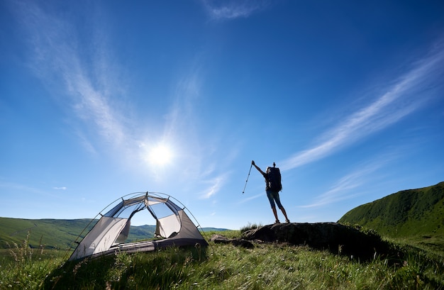 Schattenbild des Mädchenwanderers mit dem Rucksack, der auf die Oberseite eines Hügels gegen blauen Himmel, Sonne und Wolken nahe Zelt steht, Hände in der Luft mit Wanderstöcken in den Händen anhebend und genießen Sommertag