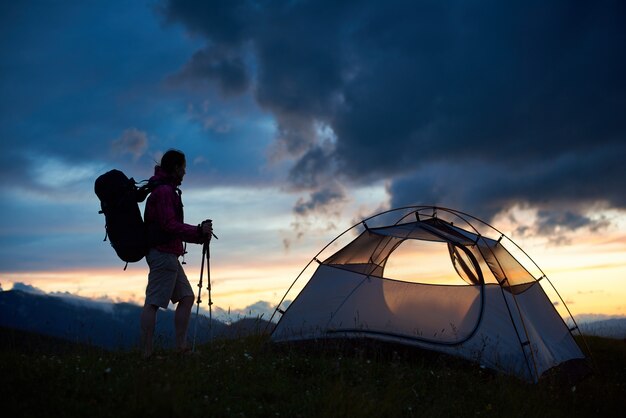 Schattenbild des weiblichen Touristen nahe dem Zelt mit Rucksack auf ihren Schultern und Gehstöcken in den Händen, die Sonnenuntergangansicht über Berghügeln genießen. Konzept für Reisen und aktive Lebensweise.