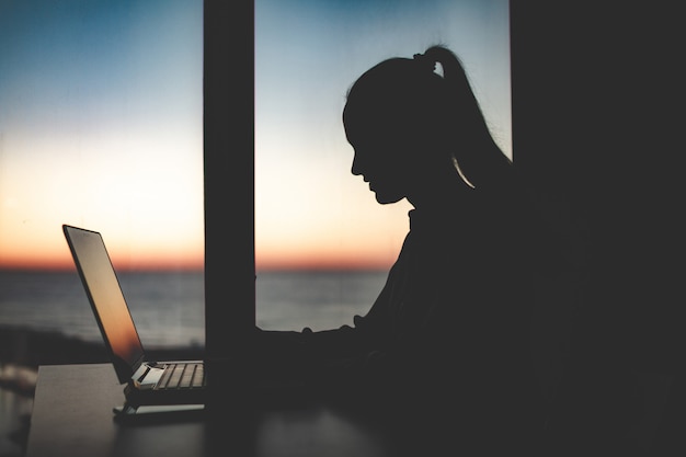 Schattenbild einer Geschäftsfrau, die spät an einem Laptop gegen das Fenster bei Sonnenuntergang arbeitet. Nacht und Abend online arbeiten