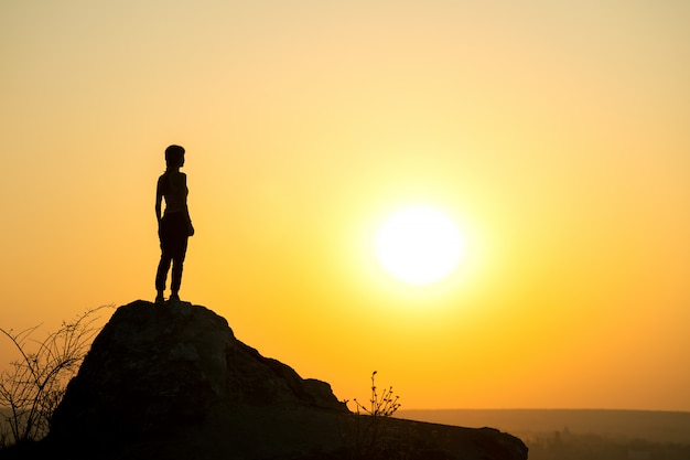 Schattenbild eines Frauenwanderers, der allein auf großem Stein bei Sonnenuntergang in den Bergen steht. Weiblicher Tourist auf hohem Felsen in der Abendnatur.