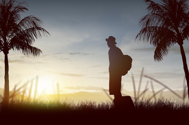 Schattenbild eines Mannes, der mit Rucksack und Koffer auf dem Feld mit Sonnenunterganghimmelhintergrund steht