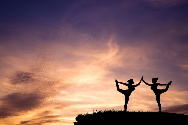 Schattenbild von zwei schönem Mädchen Yoga stehendem Bogen, der Haltung auf dem Berg mit schönem Sonnenunterganghimmel zieht.