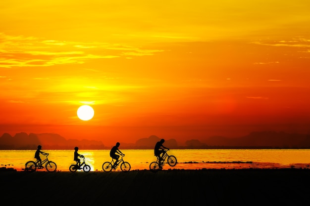 Schattenbilder von Kindern auf Fahrrad gegen Sonnenunterganghimmel am Strand.