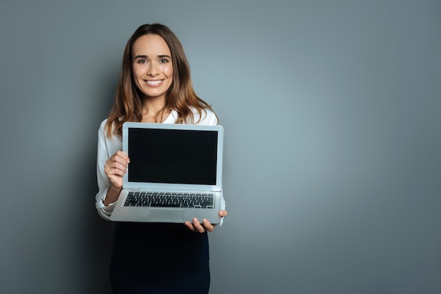 Schau auf den Bildschirm. Erfreute nette attraktive Frau, die ihren Laptop hält und lächelt, während sie es Ihnen zeigt