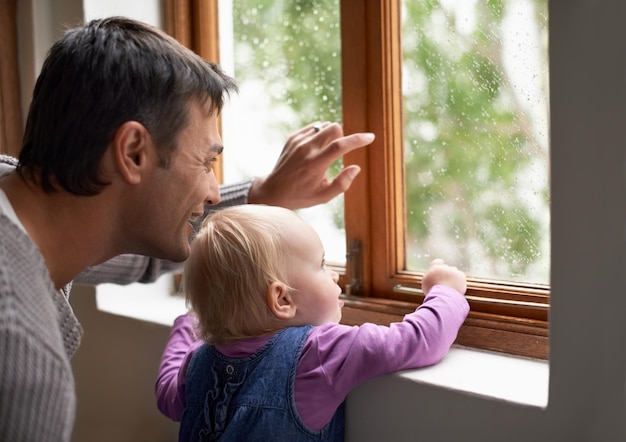 Schau dir an, was für ein schöner Tag es draußen ist Ein junger Vater und sein kleines Mädchen, die etwas hinter dem Fenster betrachten