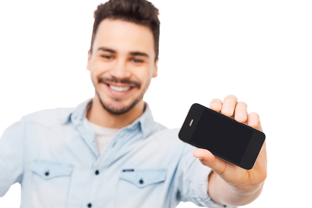 Schau dir mein neues Gadget an! Fröhlicher junger Mann, der Handy zeigt und lächelt, während er vor weißem Hintergrund steht